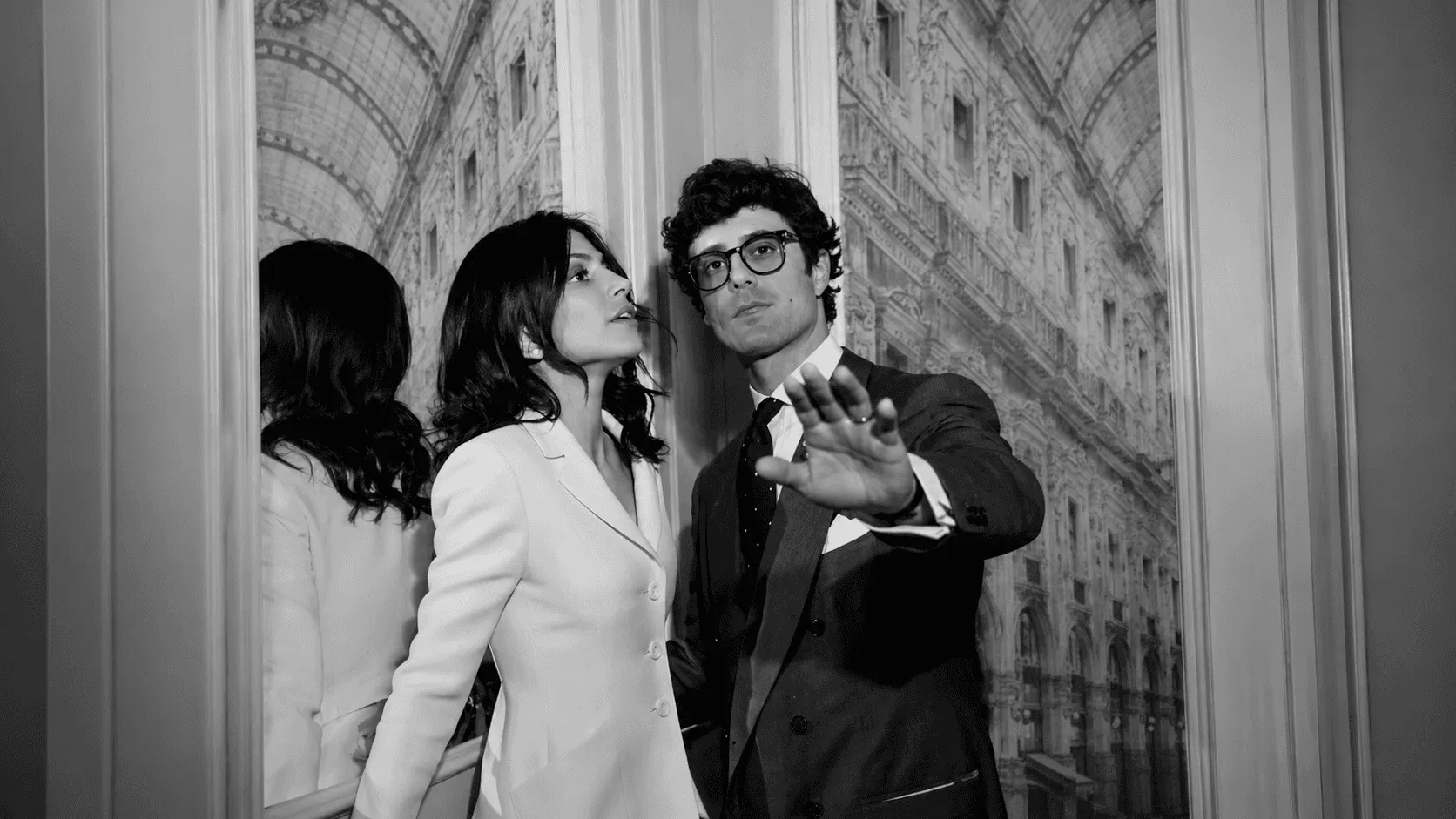 A stylishly dressed man and woman pose in front of an ornate, classical-style building facade, reflected in a large mirror, exuding elegance and sophistication in a black-and-white photograph.
