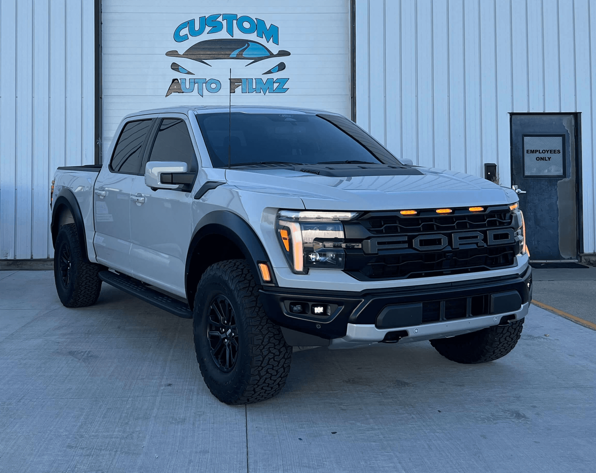 A silver Ford F-150 Raptor pickup truck is parked on a concrete driveway in front of a white industrial building.