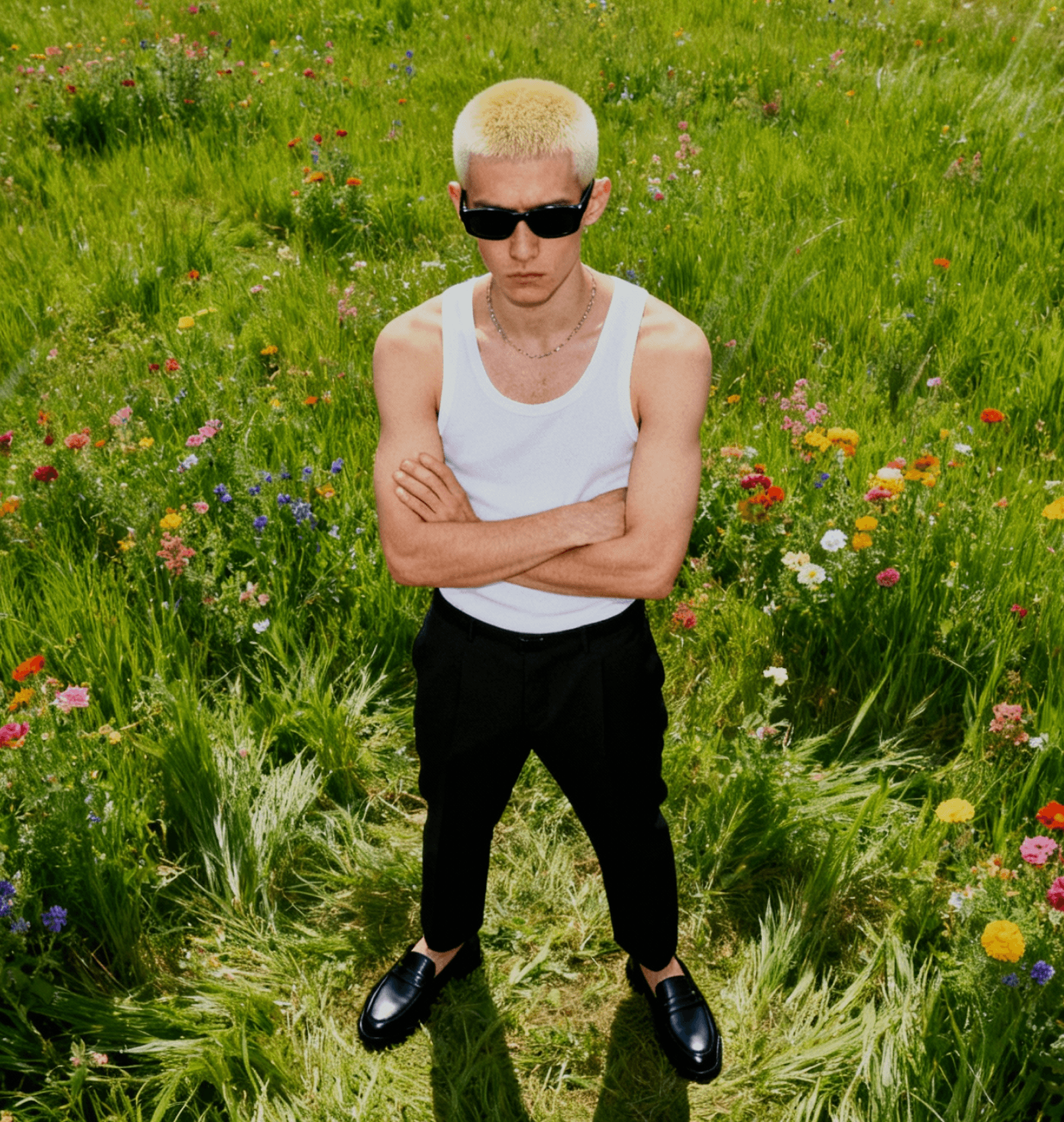 Person with platinum blonde buzz cut wearing black sunglasses, white tank top, and black pants standing with arms crossed in a wildflower meadow with colorful blooms