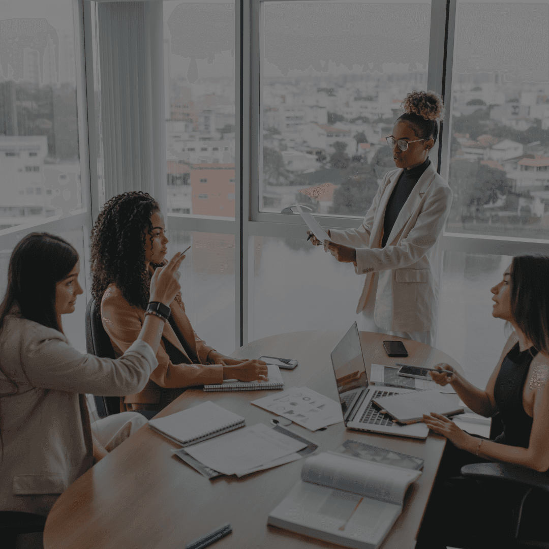 A group of professionals in a meeting, with one person presenting to others at a table. City view in the background.