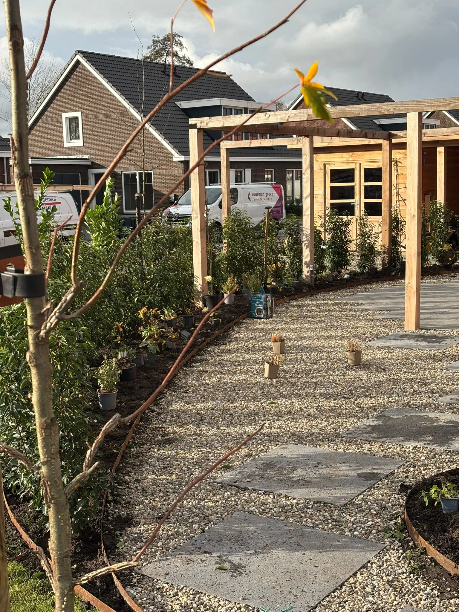 Nieuw aangelegde tuin met grindpad en grote betontegels, jonge haagplanten langs rand en houten pergola bij nieuwbouwwoning.