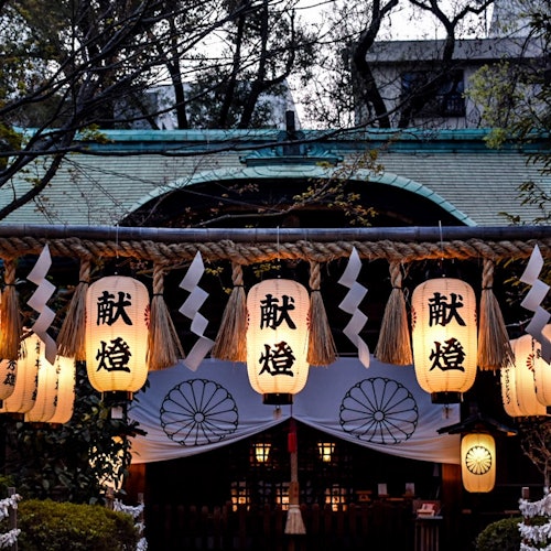 在被树木环绕的传统建筑前悬挂着带有日文字样的灯笼和白色纸质装饰。