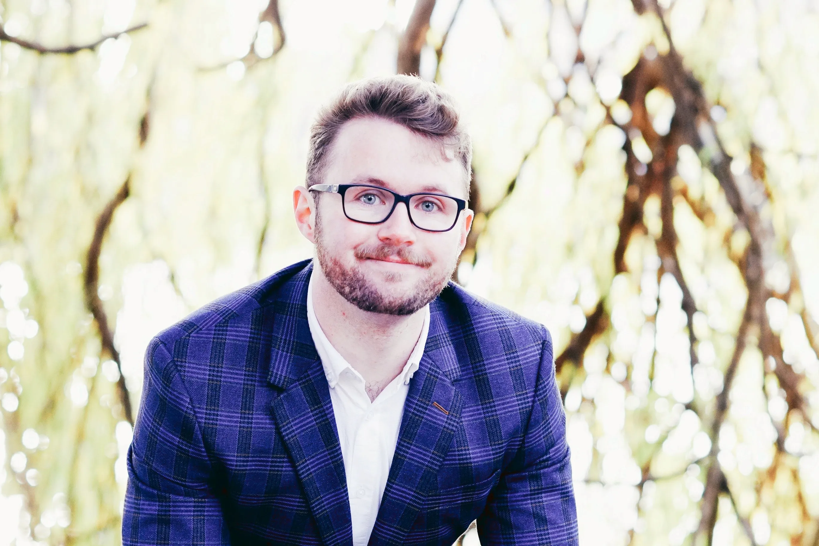 A man wearing a blue plaid suit and glasses smiles while standing in front of a backdrop of softly blurred tree branches and golden foliage.