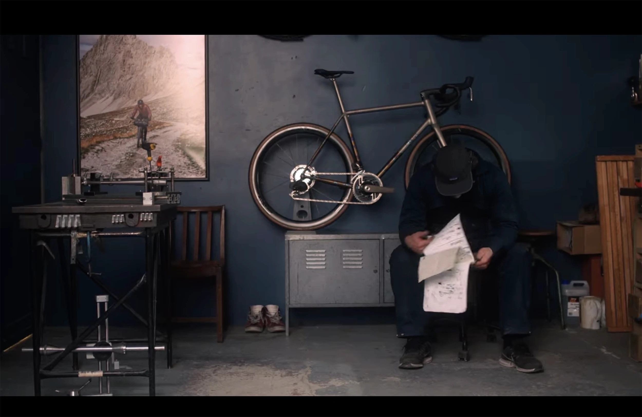 Rob Quirk sat in dark workshop in overalls, flicking though drawings. Light shines on large picture on wall and bike mounted behind him.