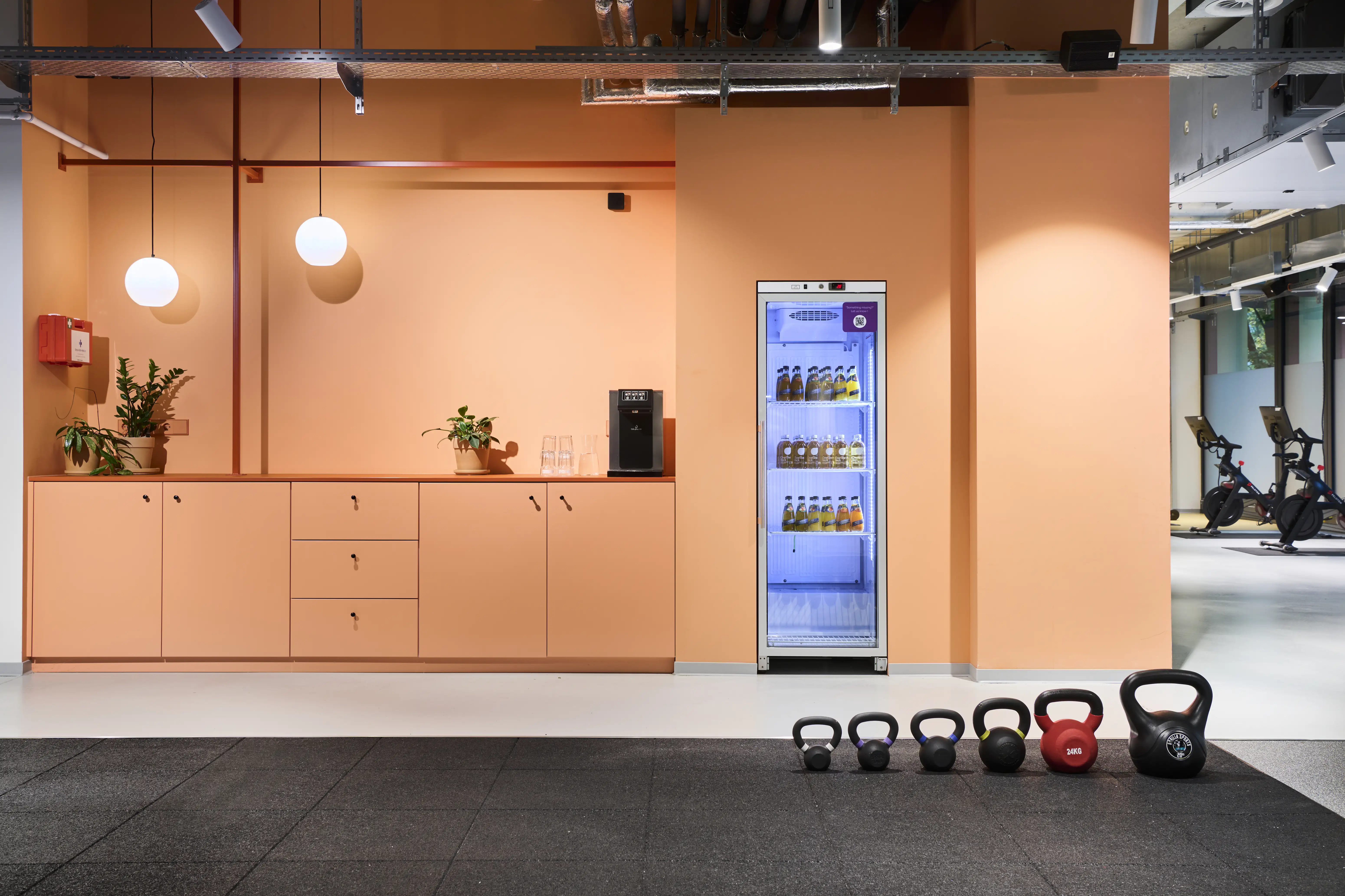 Apricot-colored kitchenette at HELLOFRESH GYM with built-in cabinets, white globe lamps, a glass-front drinks fridge and a row of kettlebells on the dark floor.