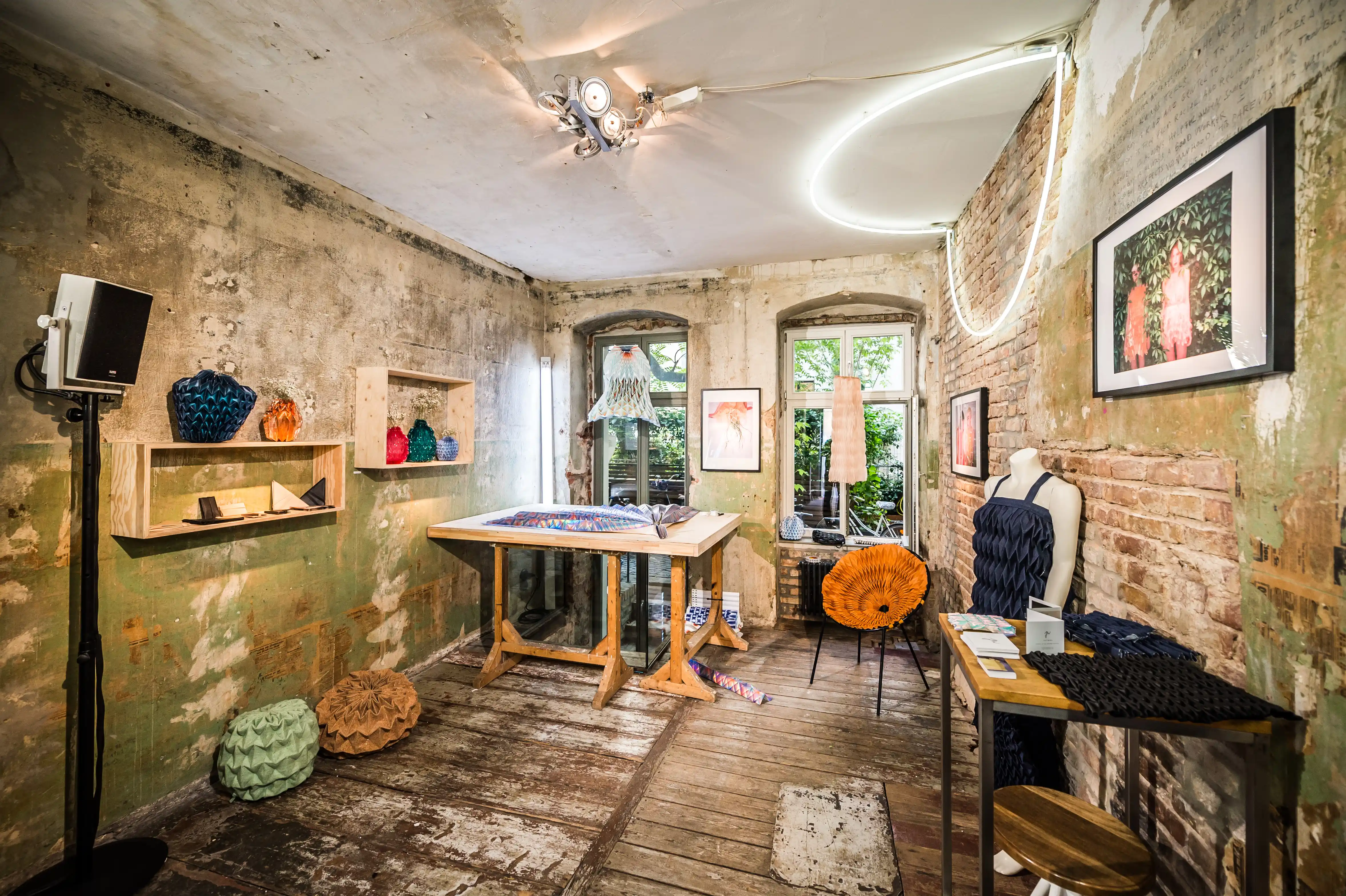 Workshop room at the MUSTANG Street ART Lab with a wooden work table, neon circle light, knitted objects on shelves and photography on raw green walls.