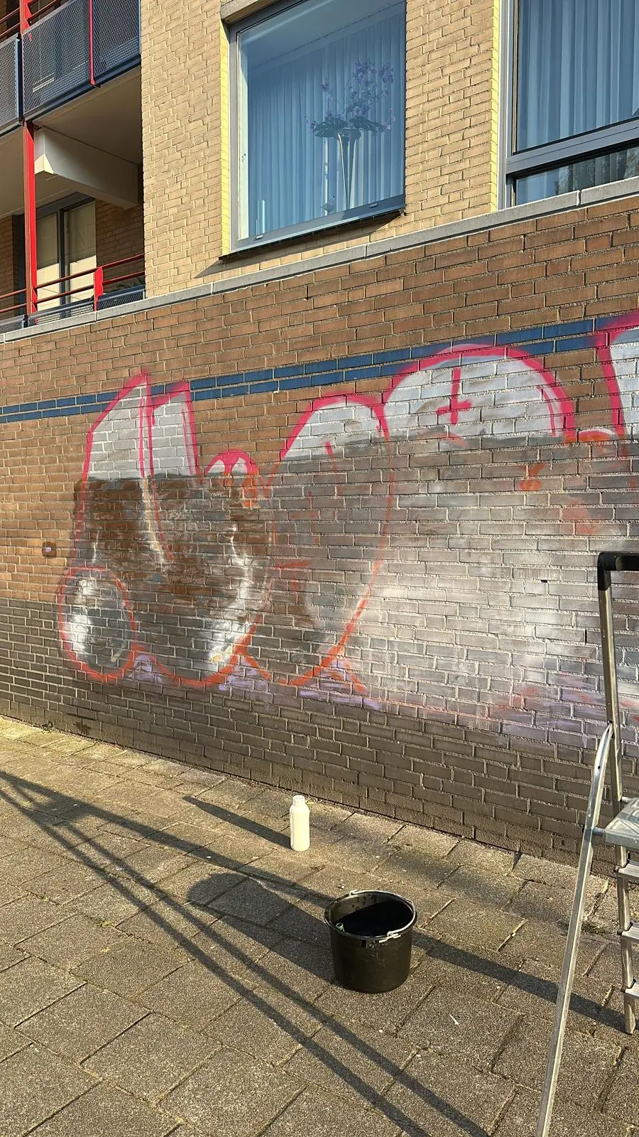 Bakstenen plintgevel van een appartementencomplex met grote roze graffitiletters die al deels behandeld zijn, met een emmer en een fles reinigingsmiddel op de stoep ervoor