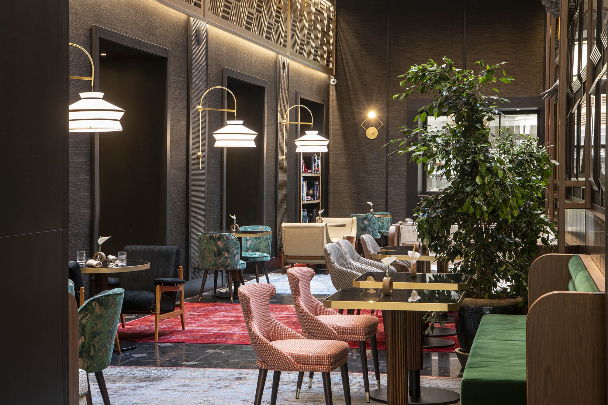 A dimly lit cafe interior with industrial decor, pendant lights, plush velvet chairs, and a large indoor tree.