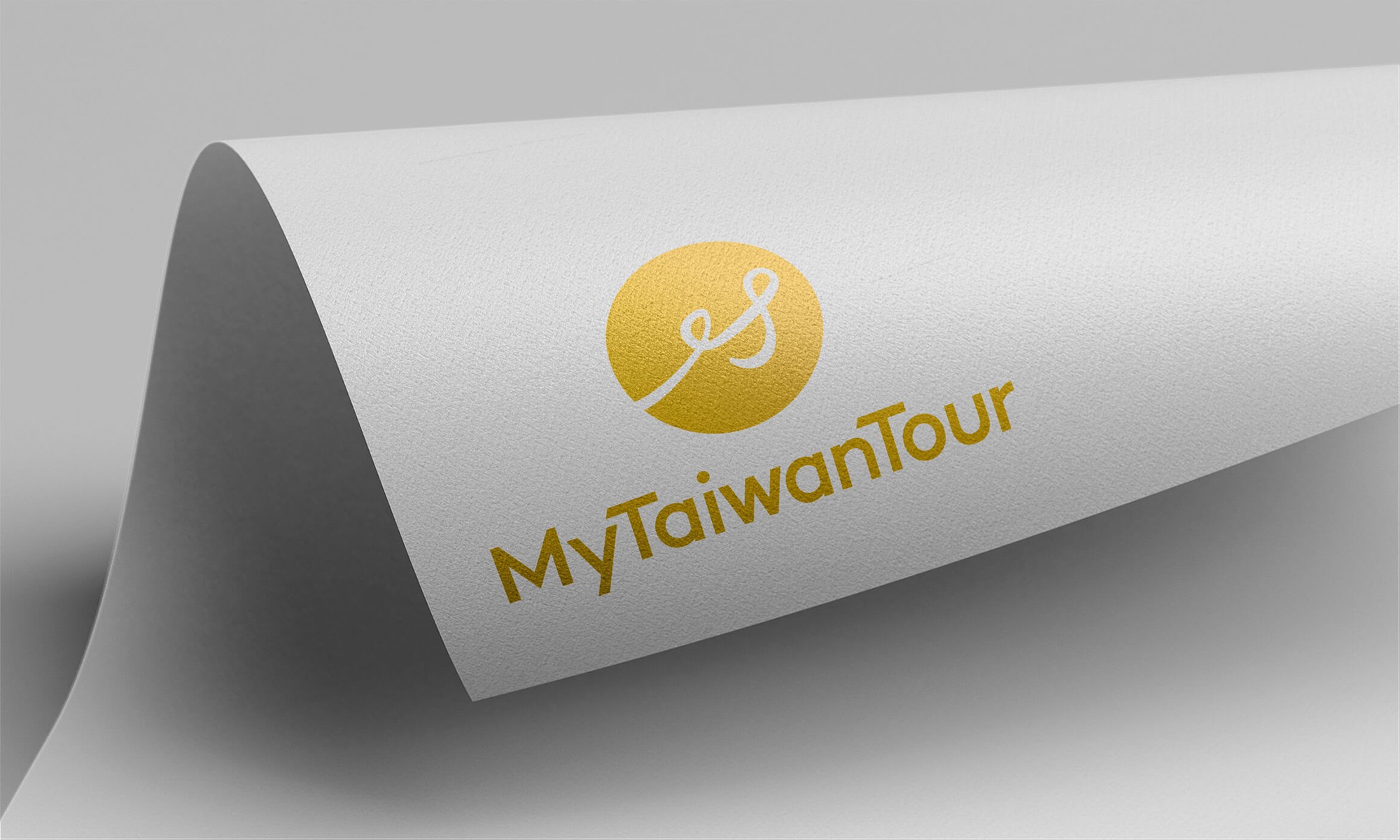 這張圖像展示了 MyTaiwanTour 的品牌識別，識別上有一個金黃色的圓形，內有白色線條構成的圖案，圓形下方有 "MyTaiwanTour" 的字樣，背景為白色紙張。