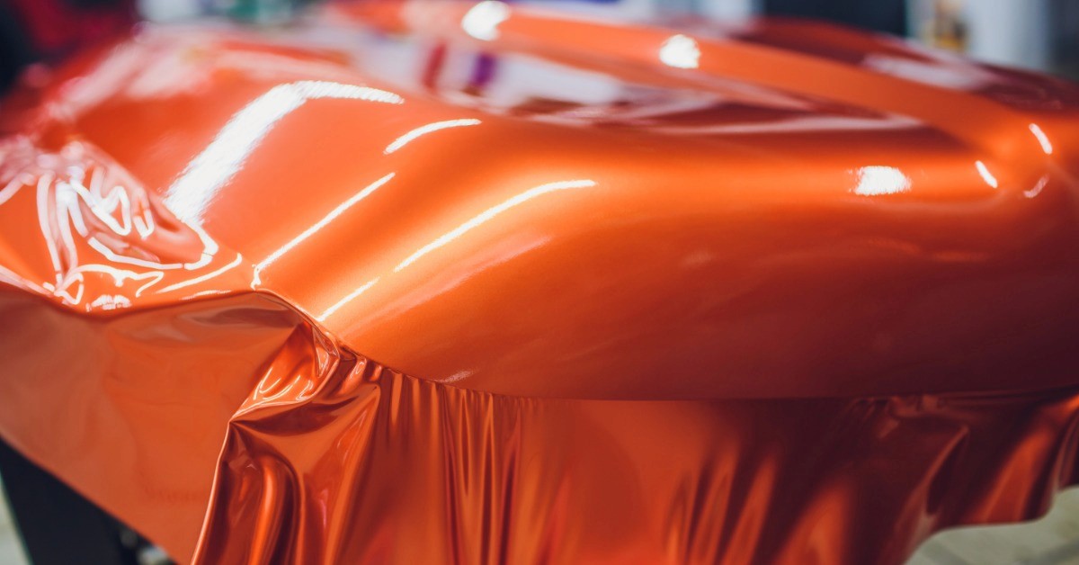 A hand pulling glossy orange vinyl film across a car body. Wrinkled material hangs along the lower edge.