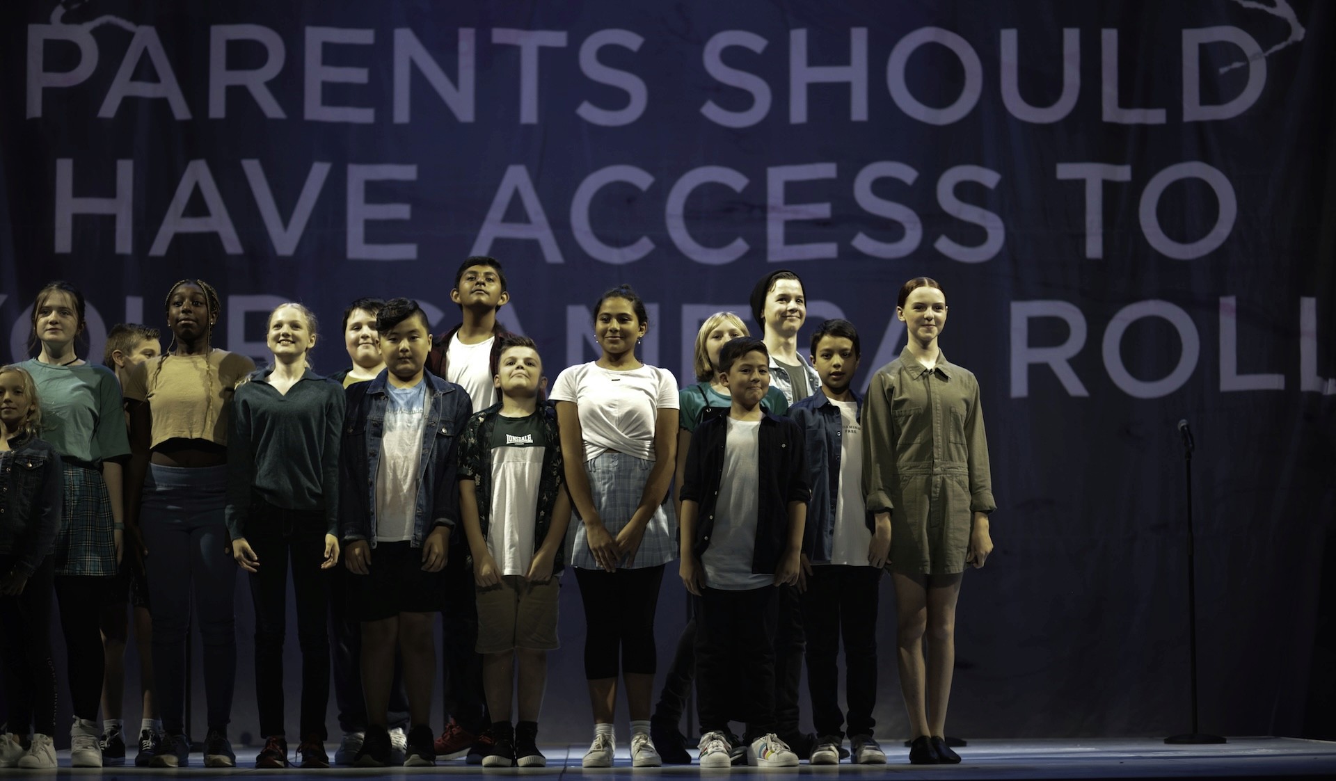 A diverse group of children and teenagers stand shoulder-to-shoulder facing the audience, lit by stage lights. Behind them, a large projection partially reads “PARENTS SHOULD HAVE ACCESS…” suggesting a bold question or statement in the performance.
