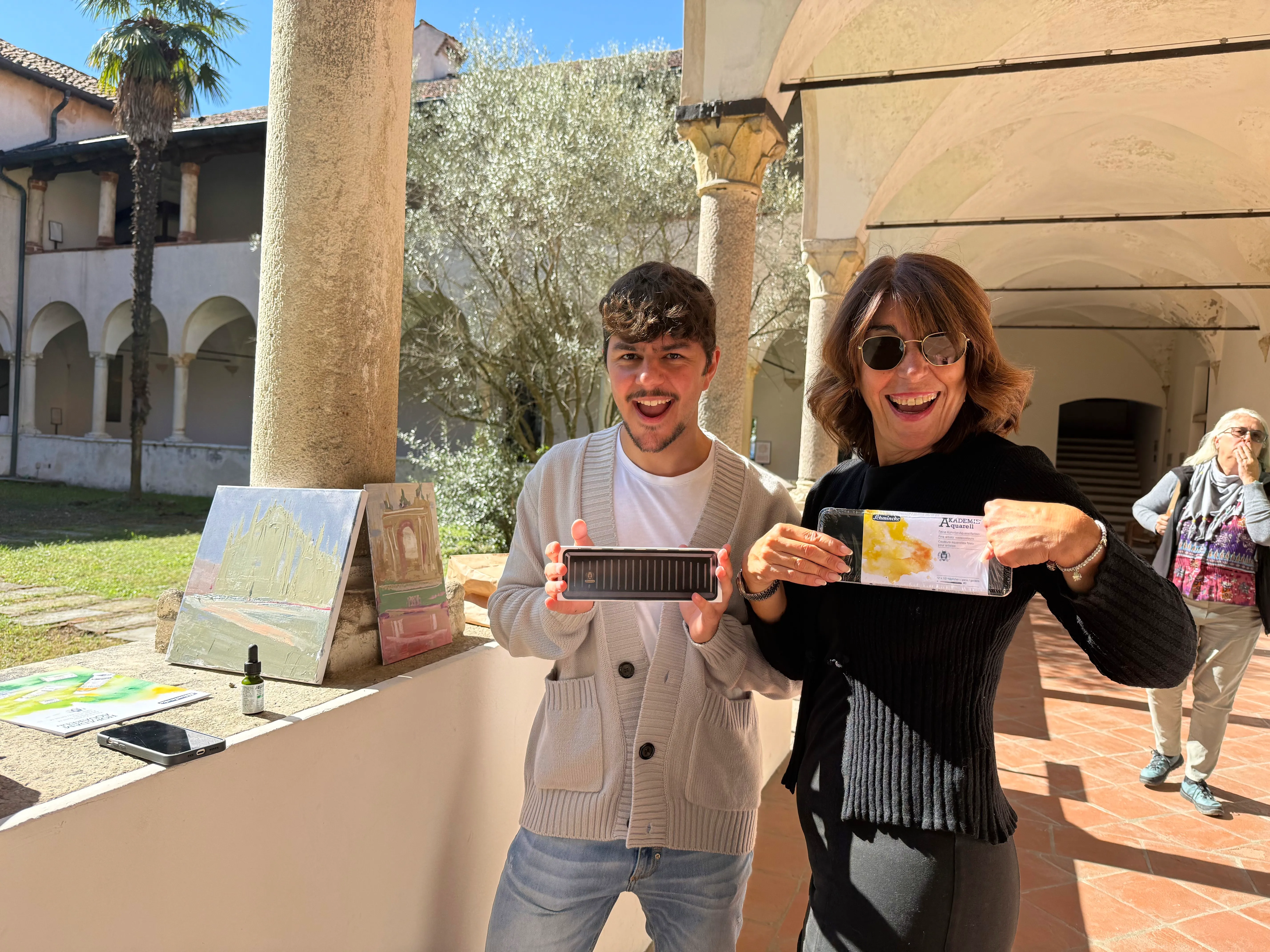 Two people smile and hold up a rectangular object and a card in a sunlit veranda with columns and artwork in the background, inside a historic building.
