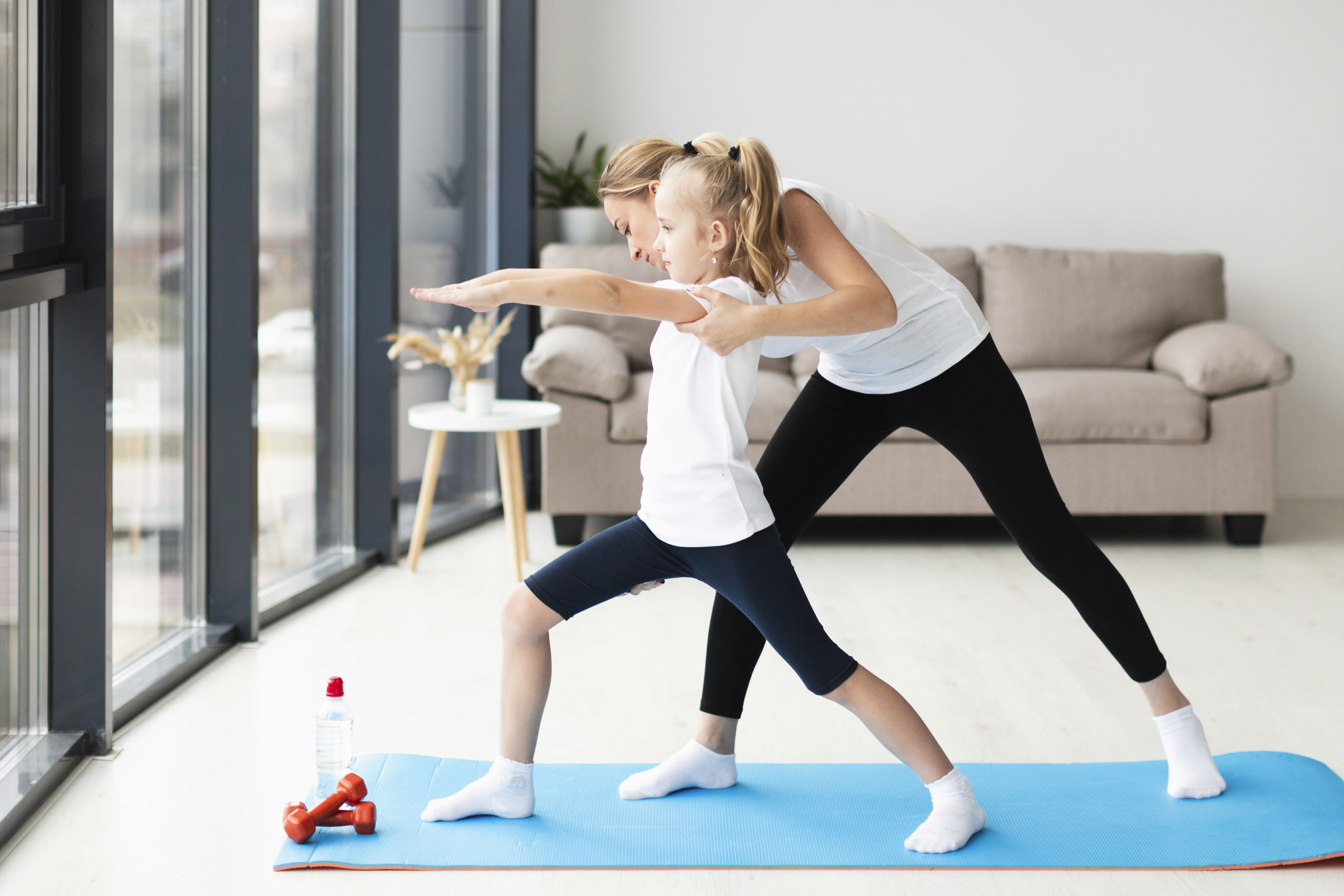 A woman helps a young girl in a yoga pose on a blue mat in a bright living room. They both look focused and calm, with a sofa and table in the background.