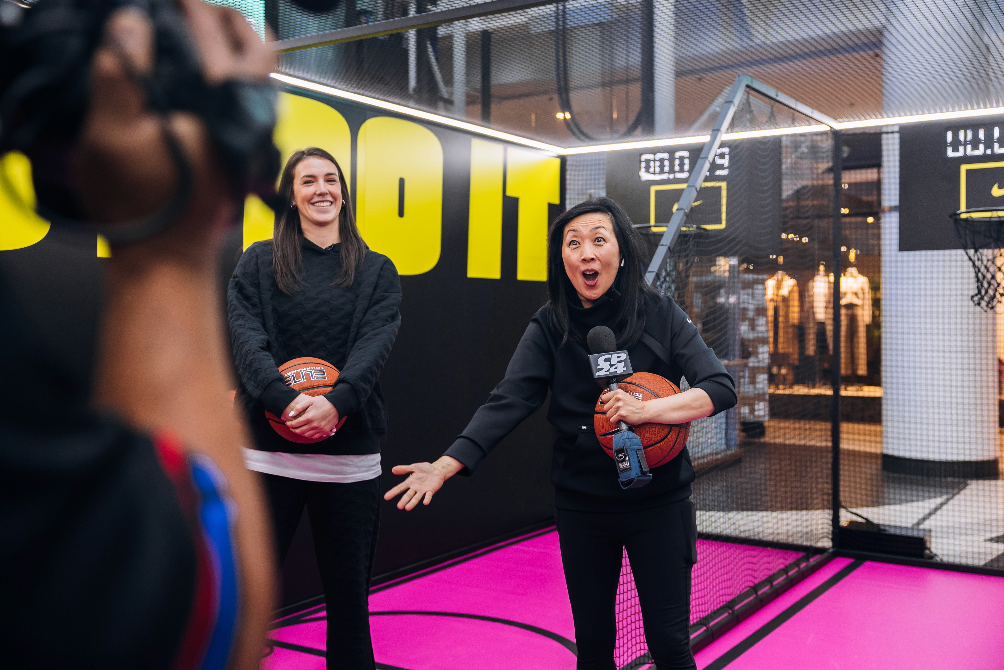 Two women are on a brightly lit indoor basketball court. One smiles while the other appears surprised, holding a microphone and basketball. Bold text reads "JUST DO IT."