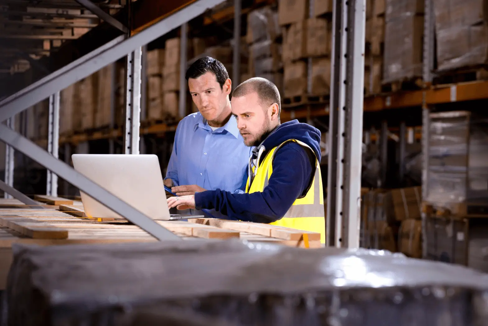 Two warehouse workers using a laptop among stacked pallets