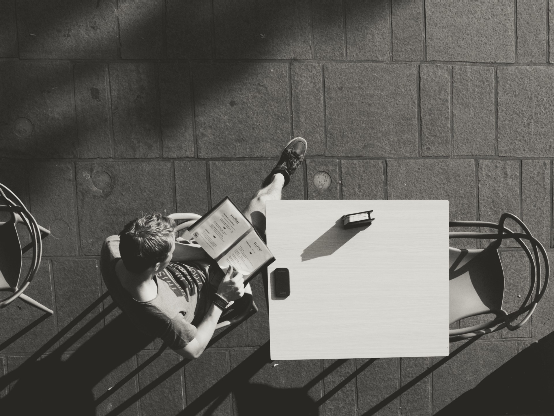 Black-and-white outdoor café photo showing a solitary person reading a menu at a sidewalk table; phone and wallet on the table, dramatic sunlit shadows on paving, ideal for street photography, minimalist lifestyle, and urban café scene content.