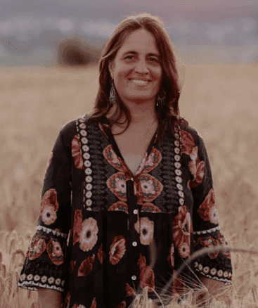 A smiling man with curly hair is sitting at a desk, looking at the camera, with a laptop and a lamp in the background.