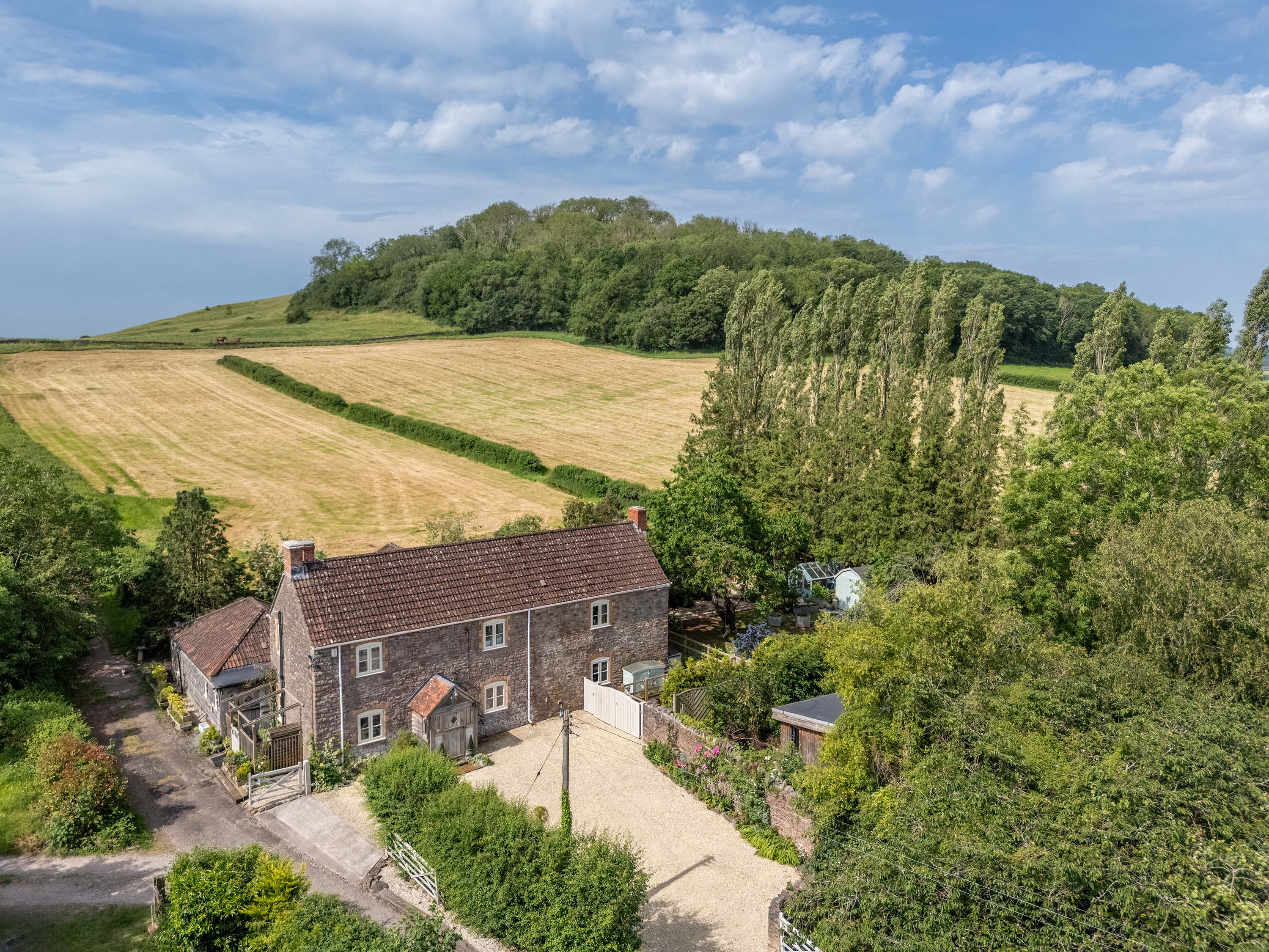 Aerial view of Rookery Farmhouse, a stone farmhouse with a clay-tile roof below fields and woodland in Nyland, near Cheddar — property photography by Captura Media