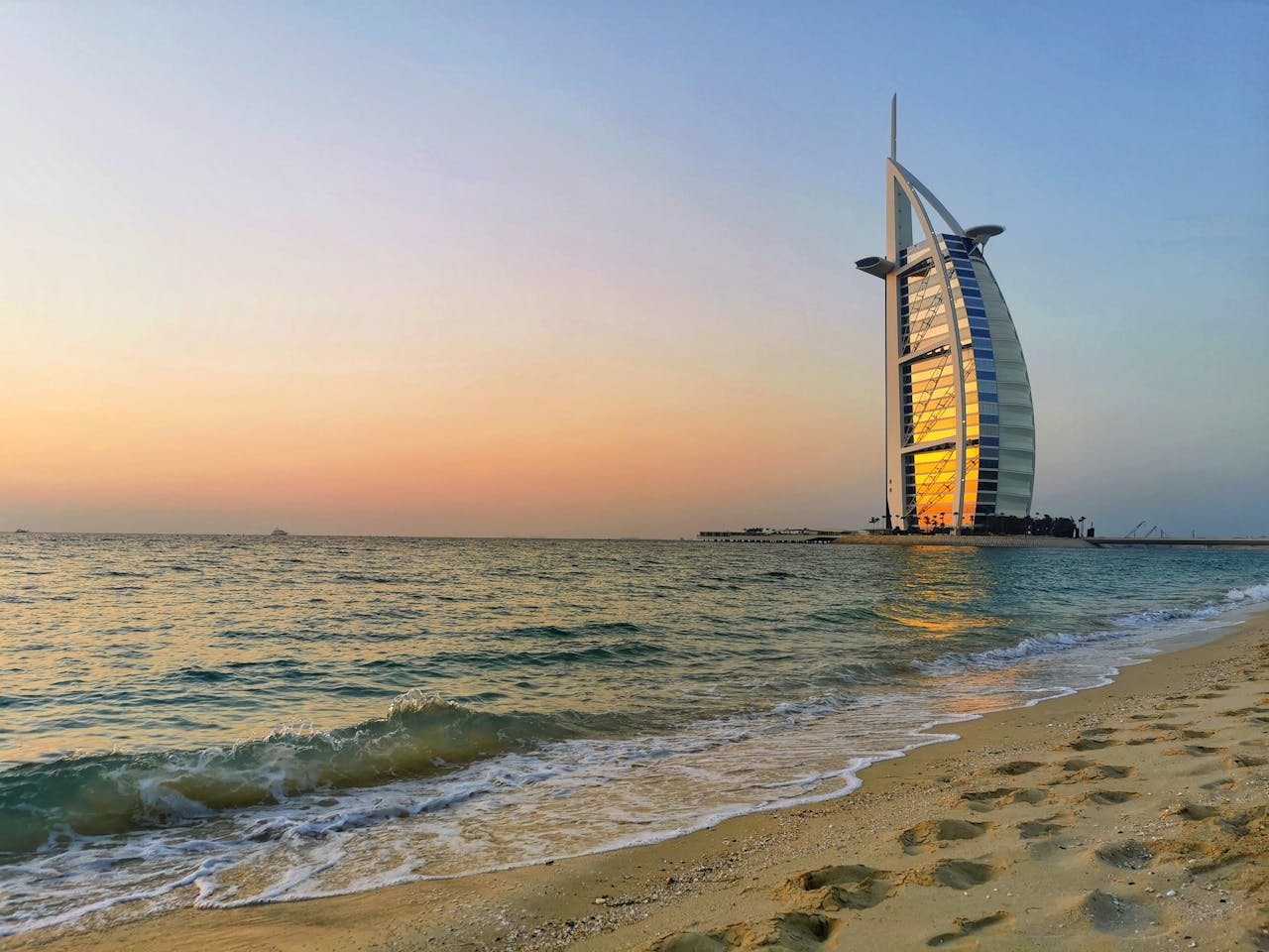 A view of the Burj Al Arab along the coastline with calm waves and the sun setting.