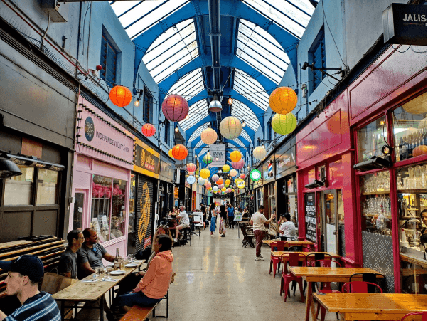 A smiling man with curly hair is sitting at a desk, looking at the camera, with a laptop and a lamp in the background.