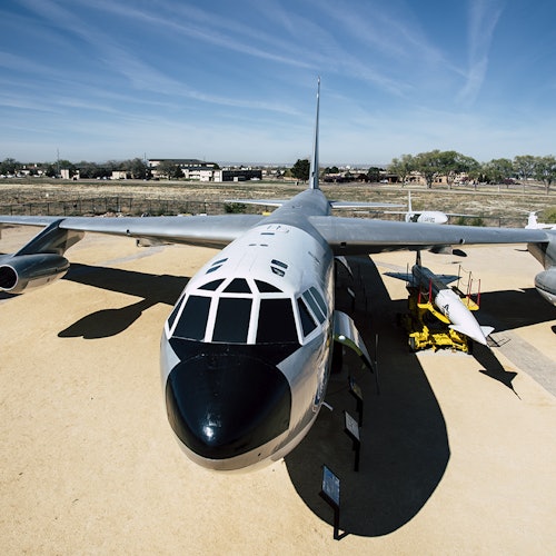 一架大型軍用飛機在戶外展示，旁邊有一架較小的飛機，背景是晴朗的天空和建築物。
