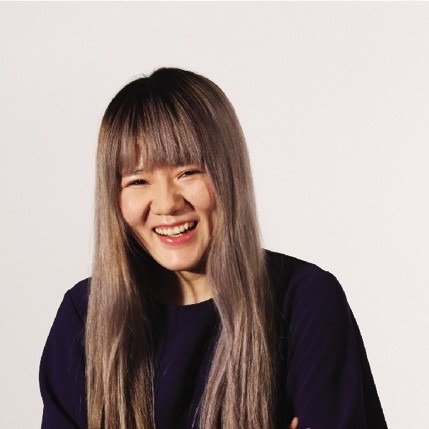 A woman with long, straight brown hair and bangs is smiling broadly, wearing a dark blue long-sleeved top. Her arms are crossed over her chest, and she stands against a plain off-white background. The lighting is even and highlights her face and hair.