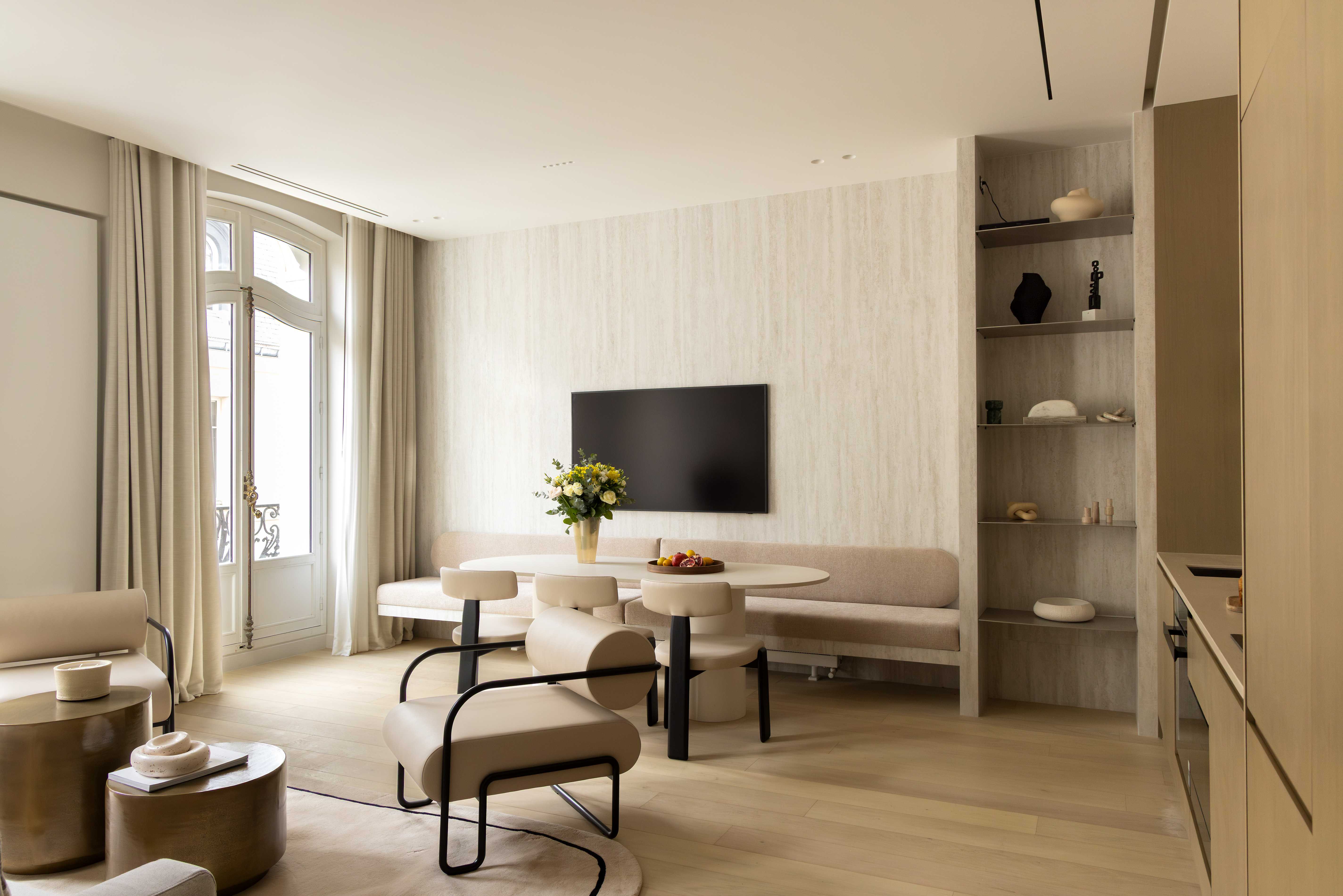 Sejour vu de biais avec banquette, table ovale, television sur mur travertin, etageres et cuisine ouverte