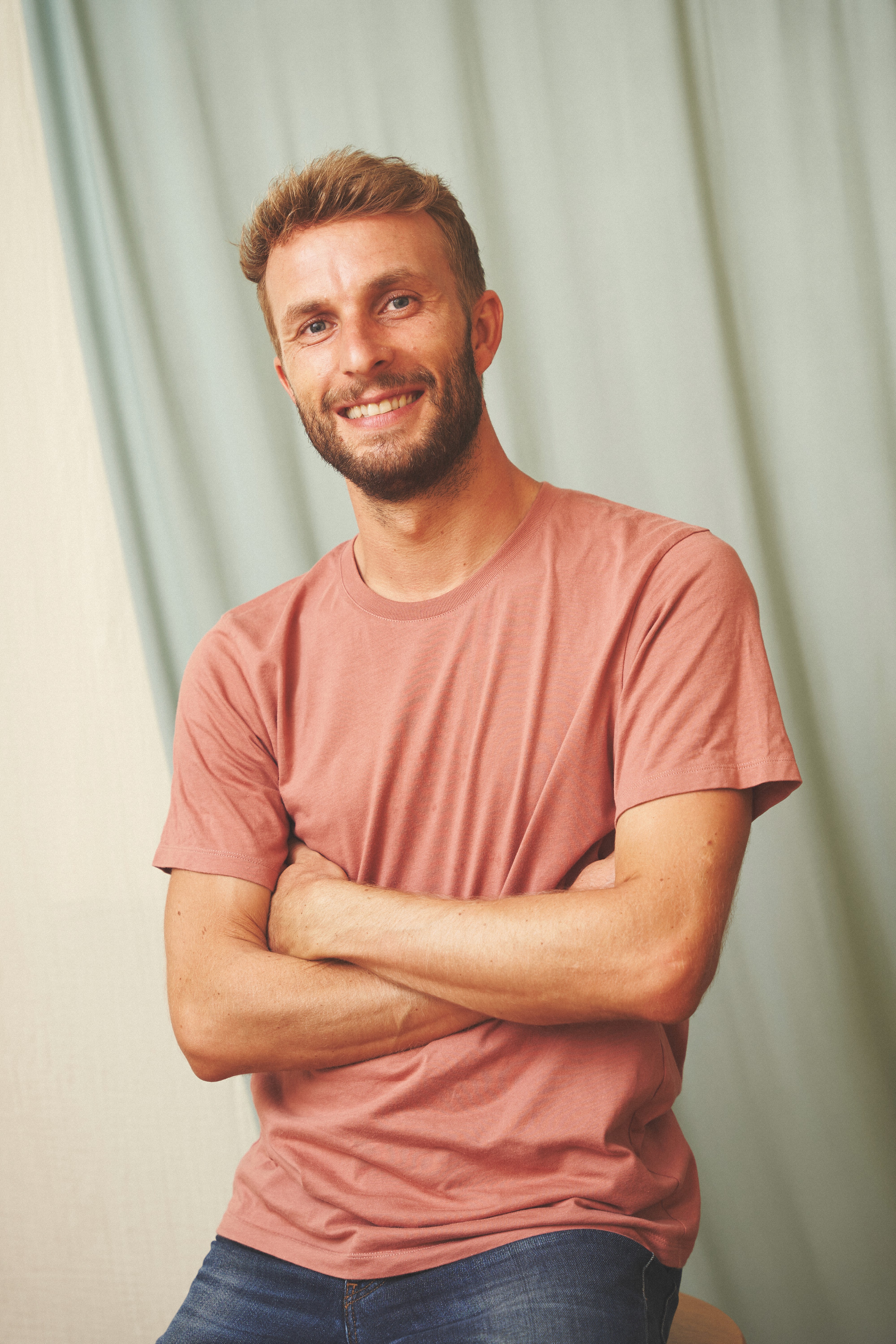 Person wearing a blue puffer jacket and red backpack standing in front of a light blue building on a city street.