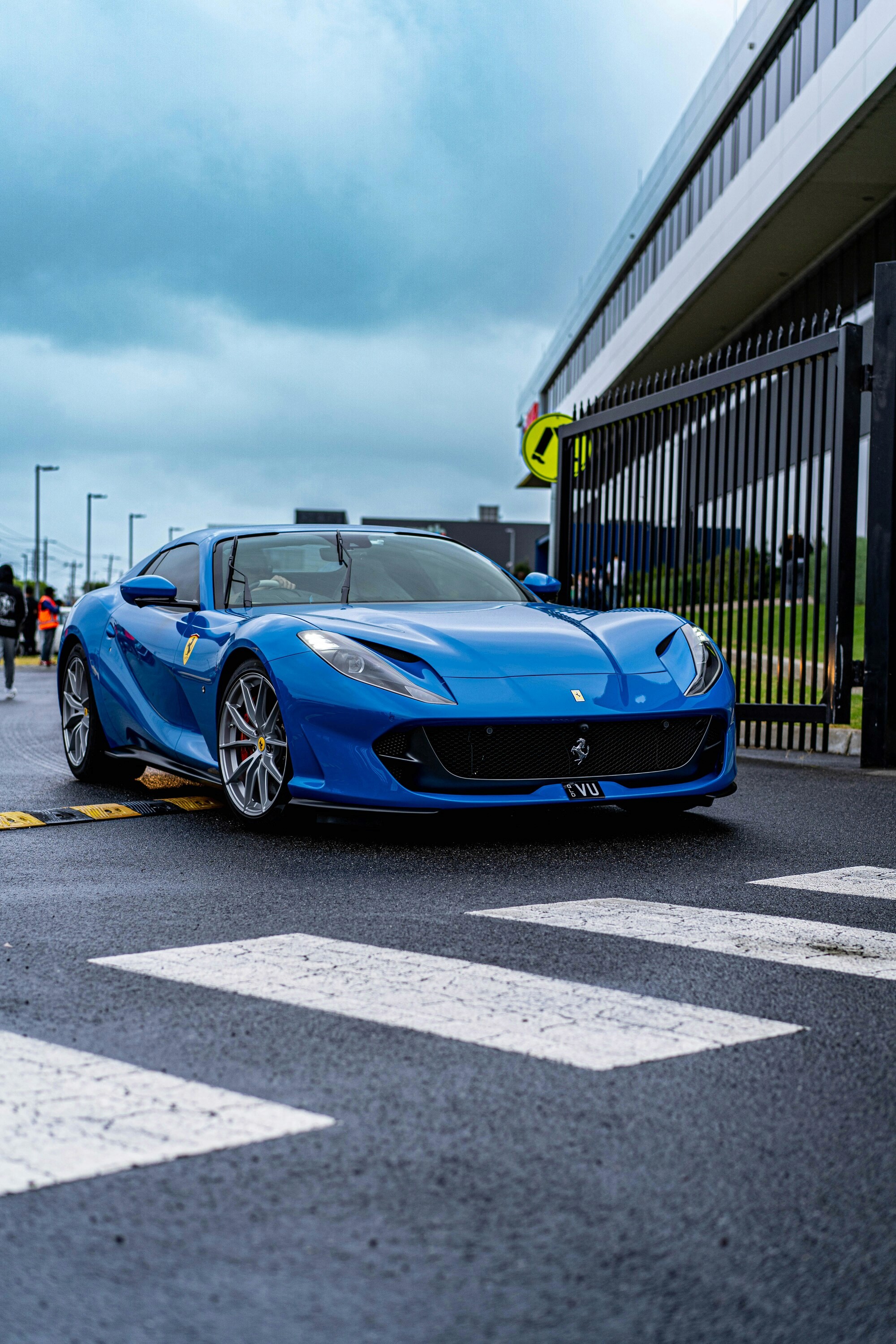 A vibrant blue sports car with sleek aerodynamic lines is parked on a wet asphalt surface by a pedestrian crossing near a metal fence and a modern building, under an overcast sky.