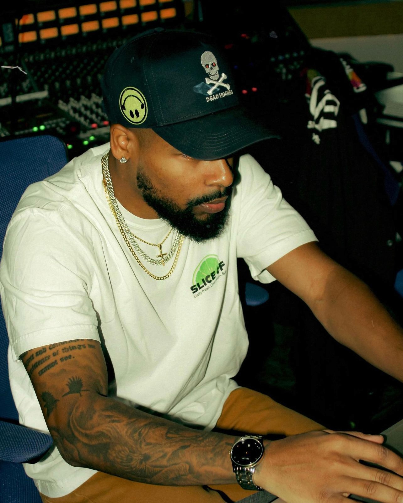 RØWAN, a rising music producer known for his work with Bryson Tiller, GIVĒON, Russ, and Rick Ross, seated in a recording studio wearing a branded cap and white t-shirt.