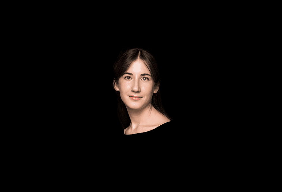 A woman with dark brown hair pulled back is centered against a solid black background. She is looking directly at the camera with a neutral expression. Her shoulders and upper chest are visible, and she wears a dark-colored top. The lighting is focused on her face, leaving the rest of the image dark and undefined.