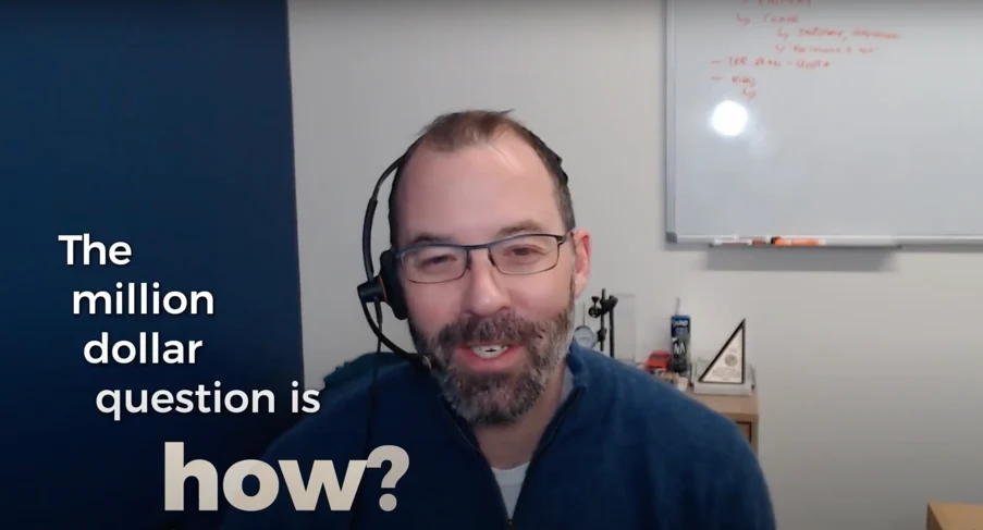 A man with a headset, smiling, sitting in front of a whiteboard, talking.