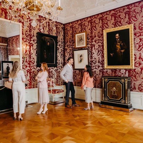 Personen in einem prächtigen Raum mit roter Damasttapete, einem kunstvollen Kronleuchter und gerahmten Porträts an den Wänden.