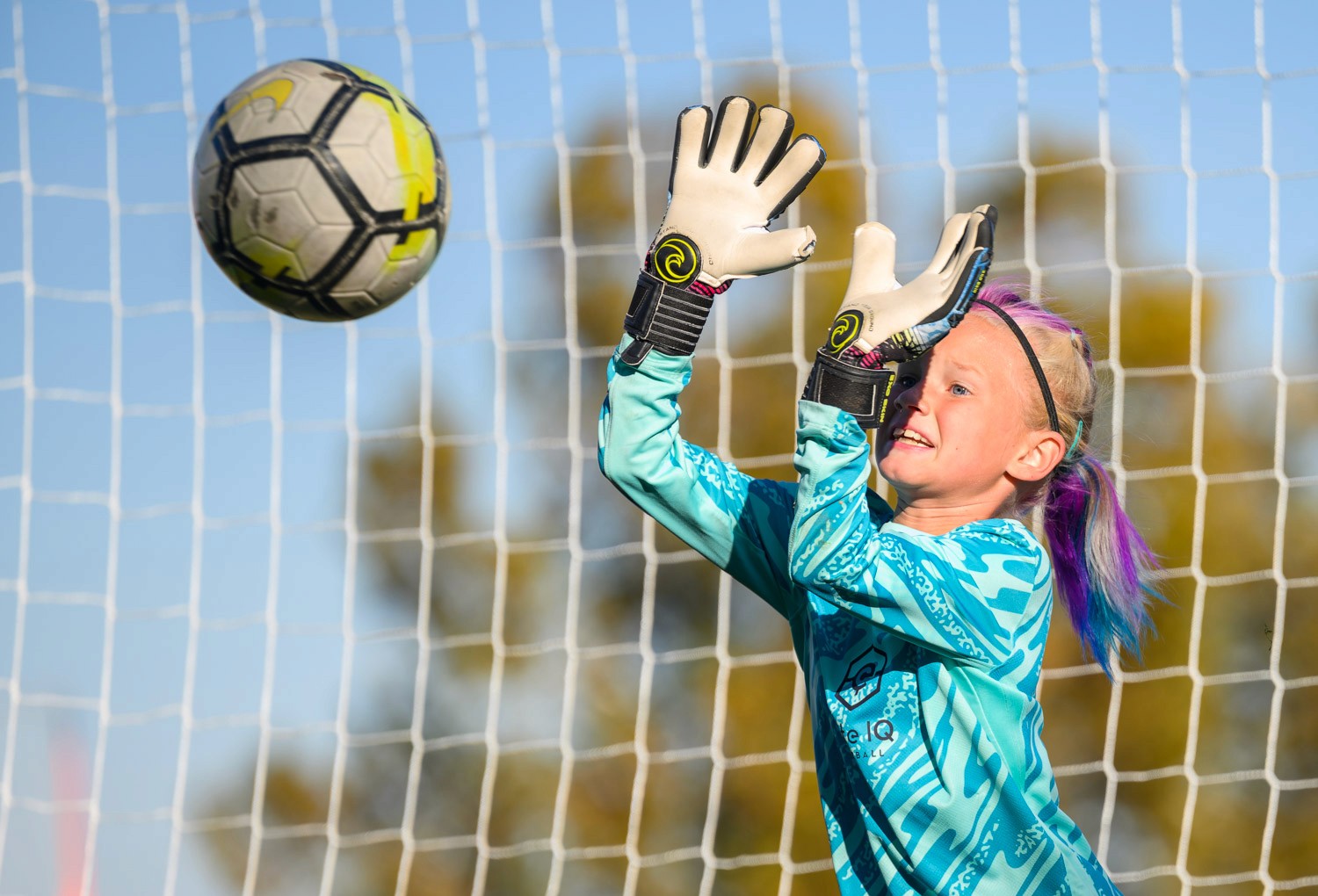 A young female goalkeeper reaches to catch a soccer ball against a bright blue sky and trees in the background.