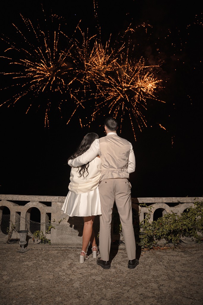 Couple seen from behind, embracing on a stone terrace while watching golden fireworks at night