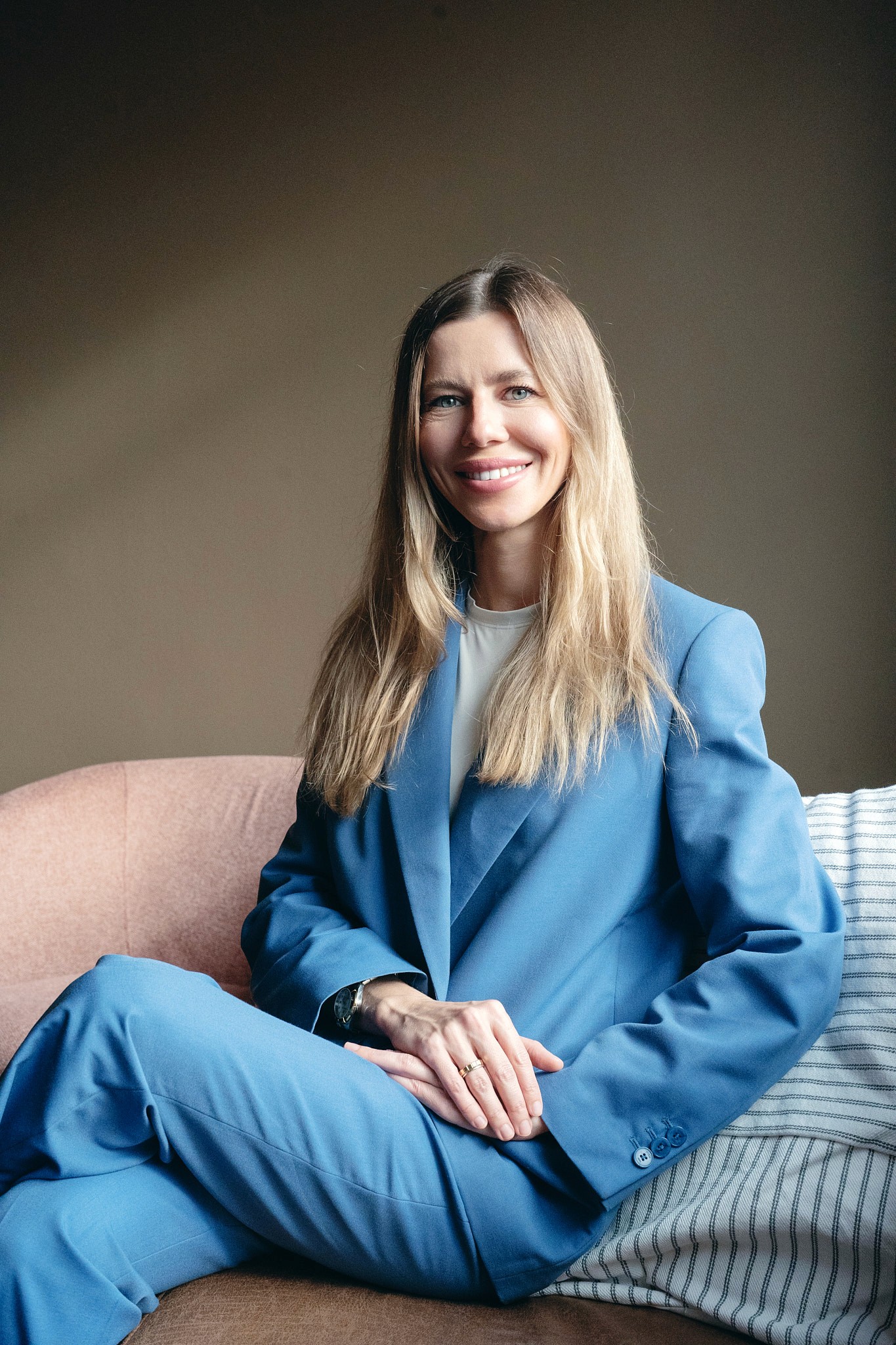 Woman in a blue suit seated on a sofa, smiling at the camera, neutral wall background.