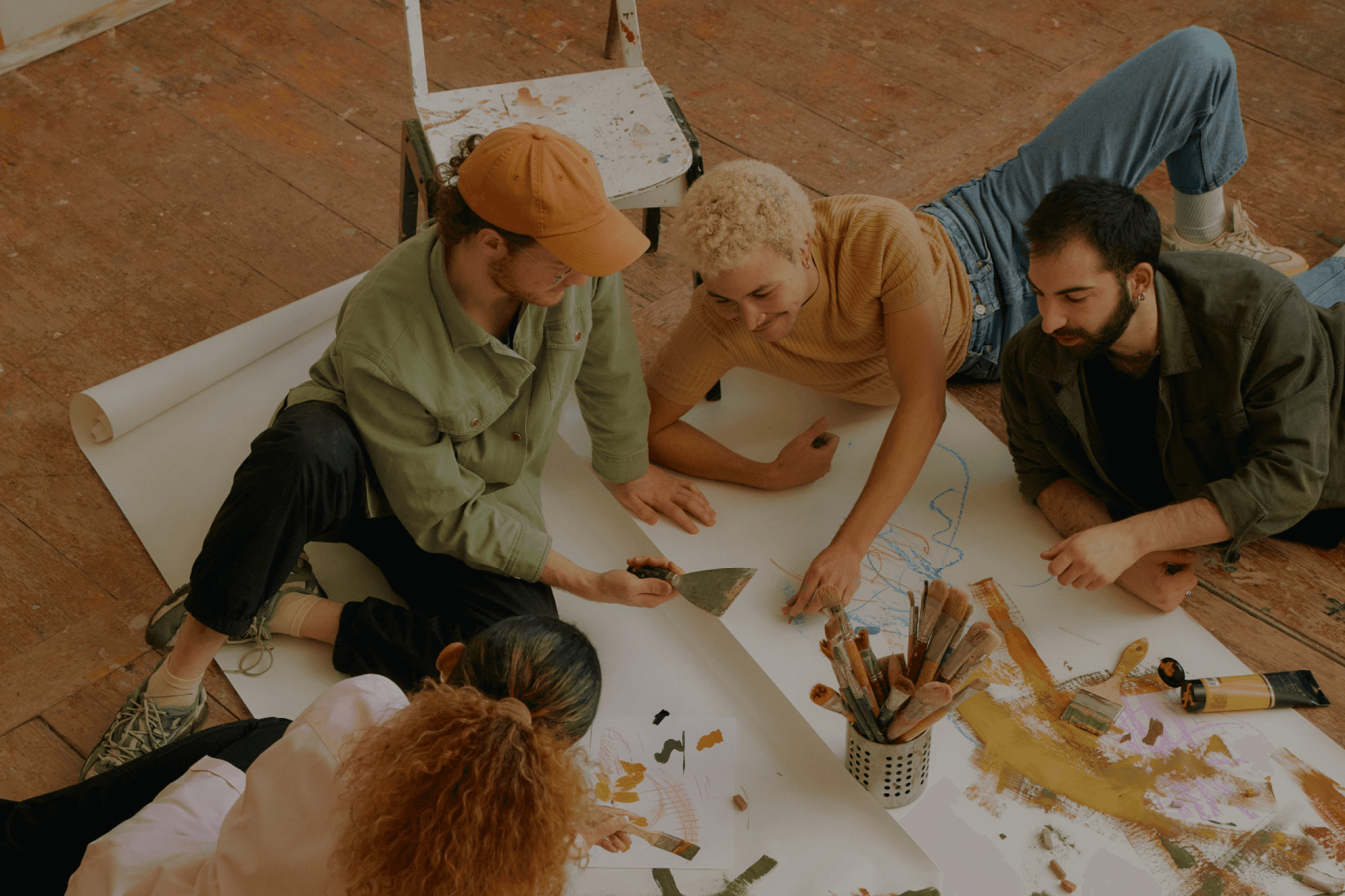 Five people lying and sitting around a long roll of paper on a studio floor, painting together.
