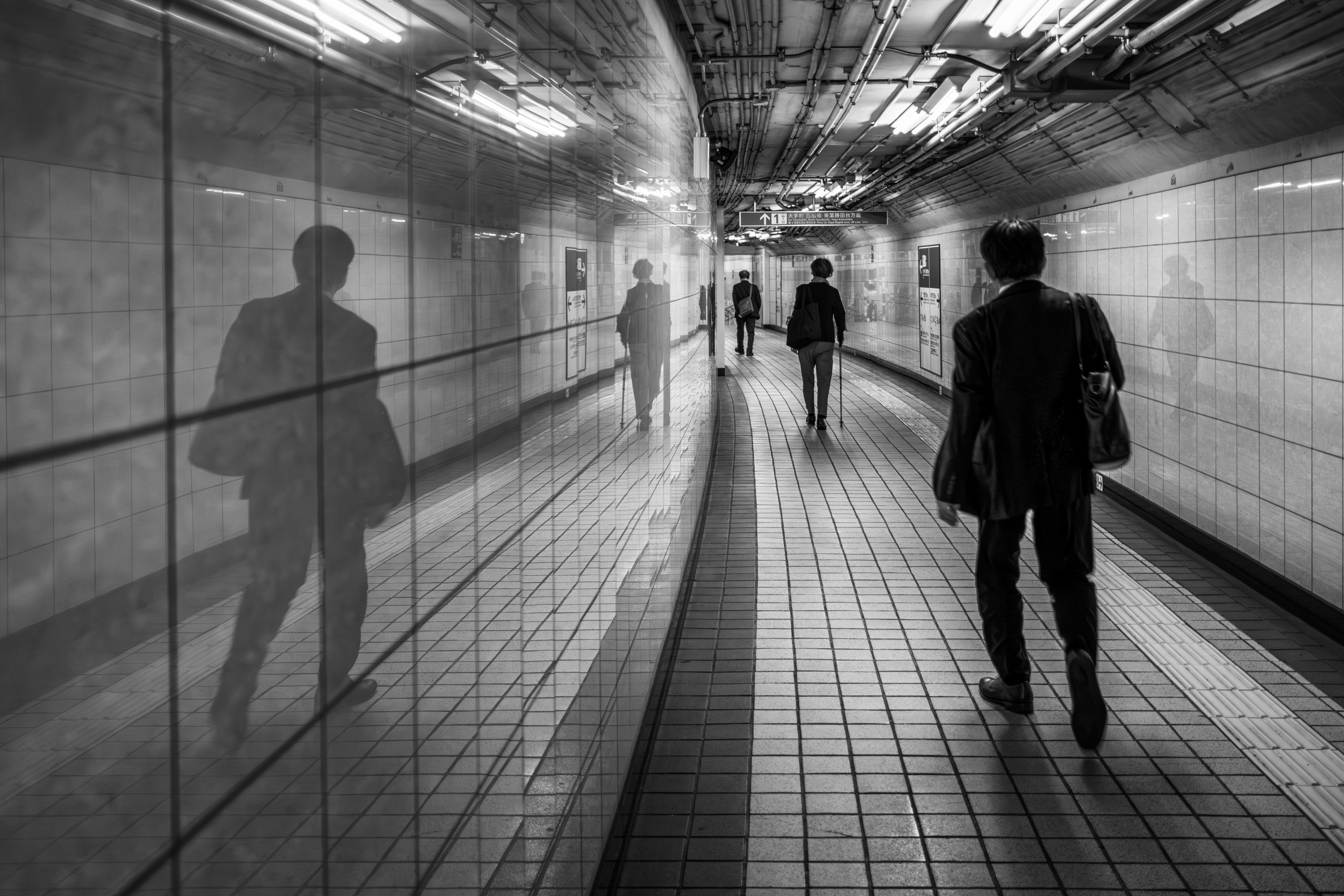 Black and white photograph of people walking through a tiled underground passageway with strong reflections.
