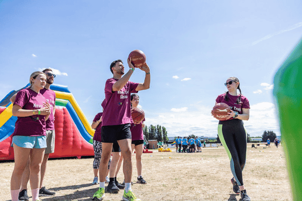 Outdoor team-building event with participants wearing matching blue shirts, taking part in inflatable obstacle courses and games, smiling and cheering, with the words 'ENTER A TEAM' in large bold white and green text across the image.