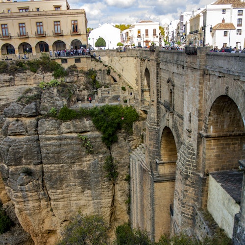Historiallinen kivisilta ylittää syvän kallioisen rotkon, yhdistäen kaksi kaupunginosaa, joissa on rakennuksia ja ihmisiä, jotka tarkkailevat ylhäältä.