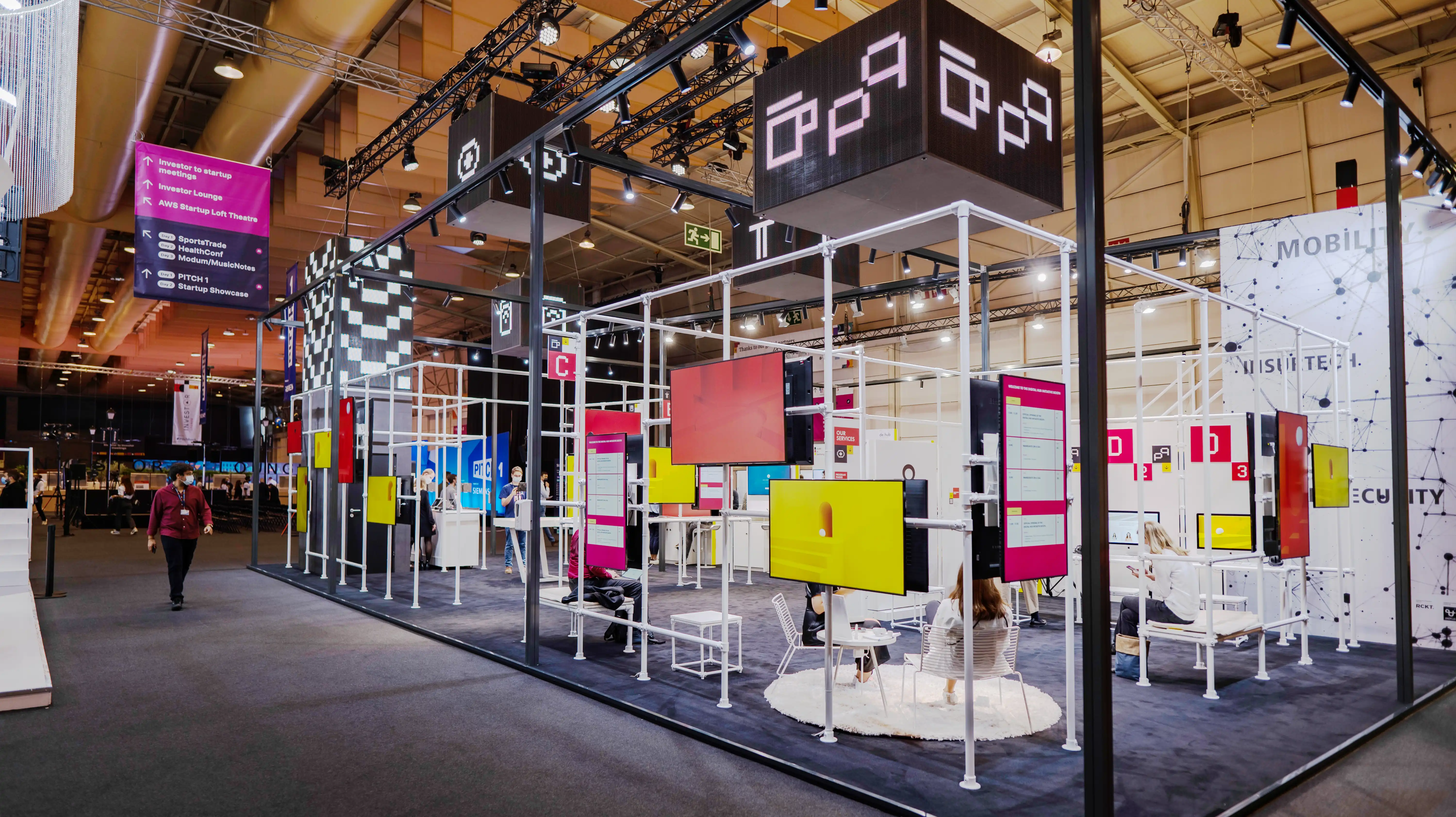 German Pavilion at Web Summit with white scaffold grid, magenta info panels and black cubes with pixel logos overhead.