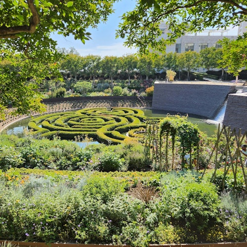 A formal garden with intricate hedges forming a spiral maze, surrounded by lush greenery and a waterfall in the background.