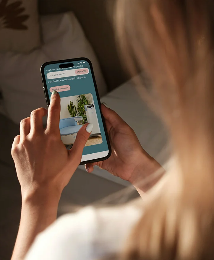 A woman sits indoors and uses her smartphone to browse the Lift and Revive website, viewing a page about continence and pelvic health treatments.