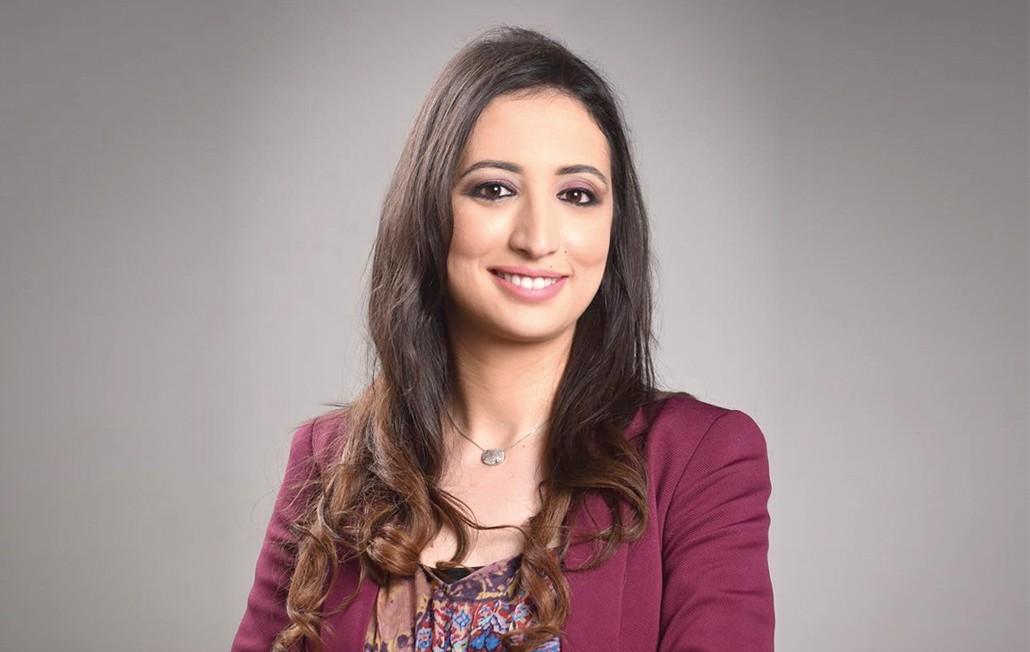 Smiling female therapist with short gray hair, wearing a brown top and a purple shawl, surrounded by a cozy background.