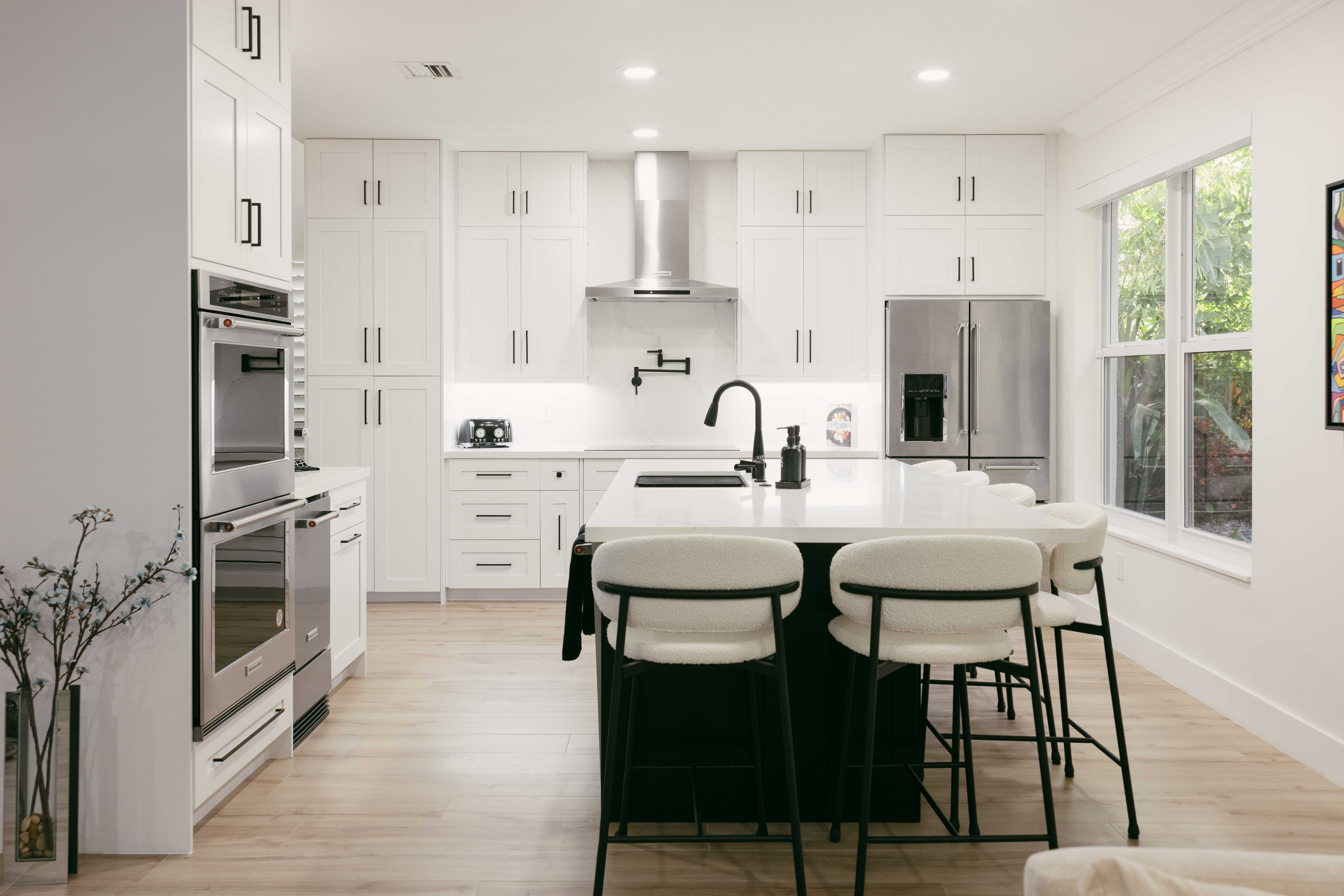 Modern white kitchen with island, stainless steel appliances, and large window.