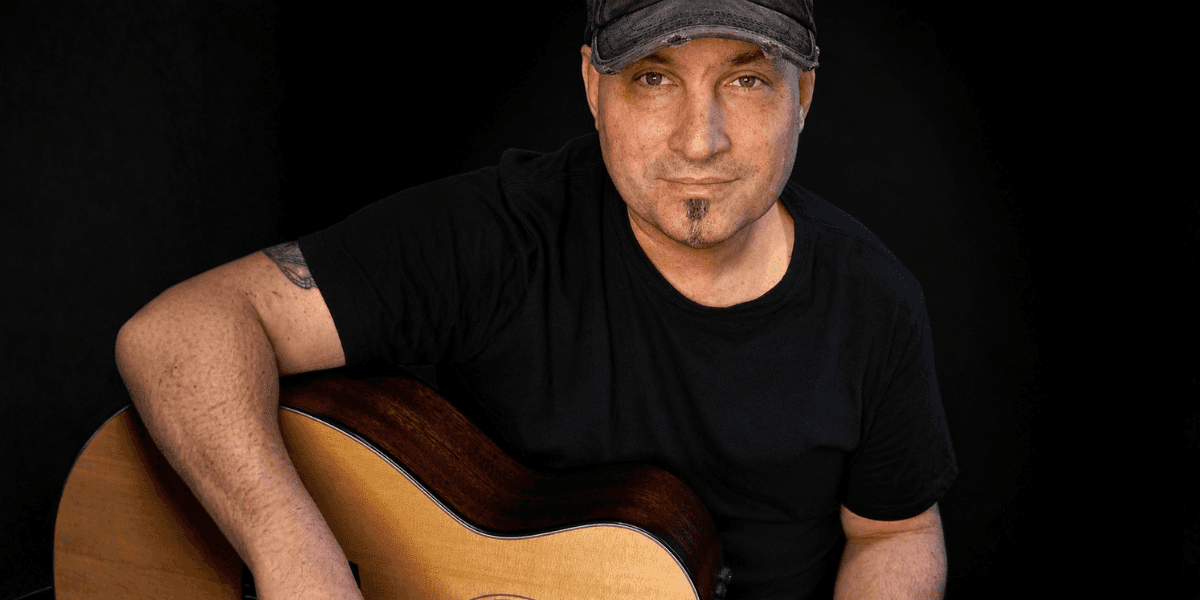 Man wearing black shirt and cap holding acoustic guitar against dark background.