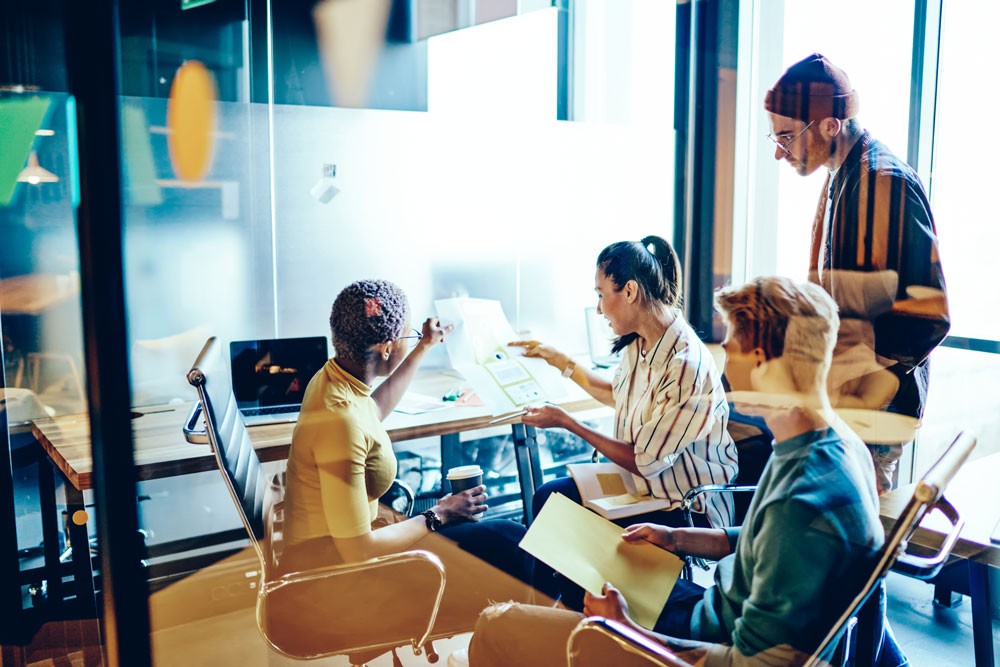 A team of young professionals collaborating in a glass-walled meeting room, reviewing documents and discussing ideas with laptops and notes on the table.