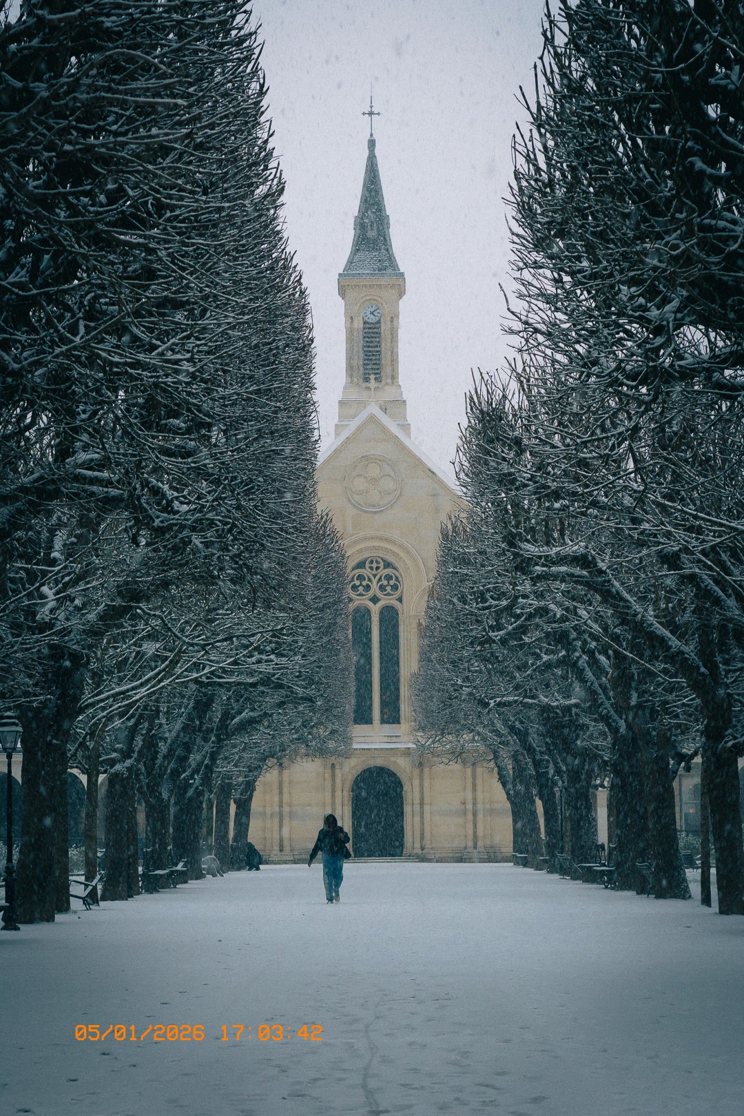 Vue hivernale de l'église Maronite Saints-Sauveur-et-Maroun sous la neige, avec un chemin bordé d'arbres enneigés et une personne marchant dans un jardin.
