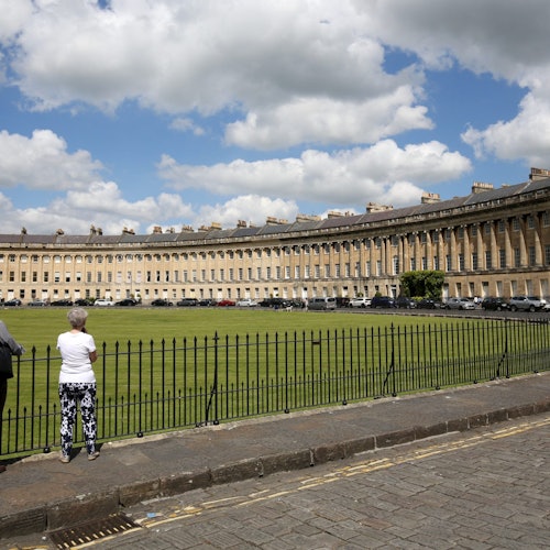 两个人站在黑色铁栅栏前,面朝一排大型弯曲的米色阶梯住宅,天空中飘着几朵白云。