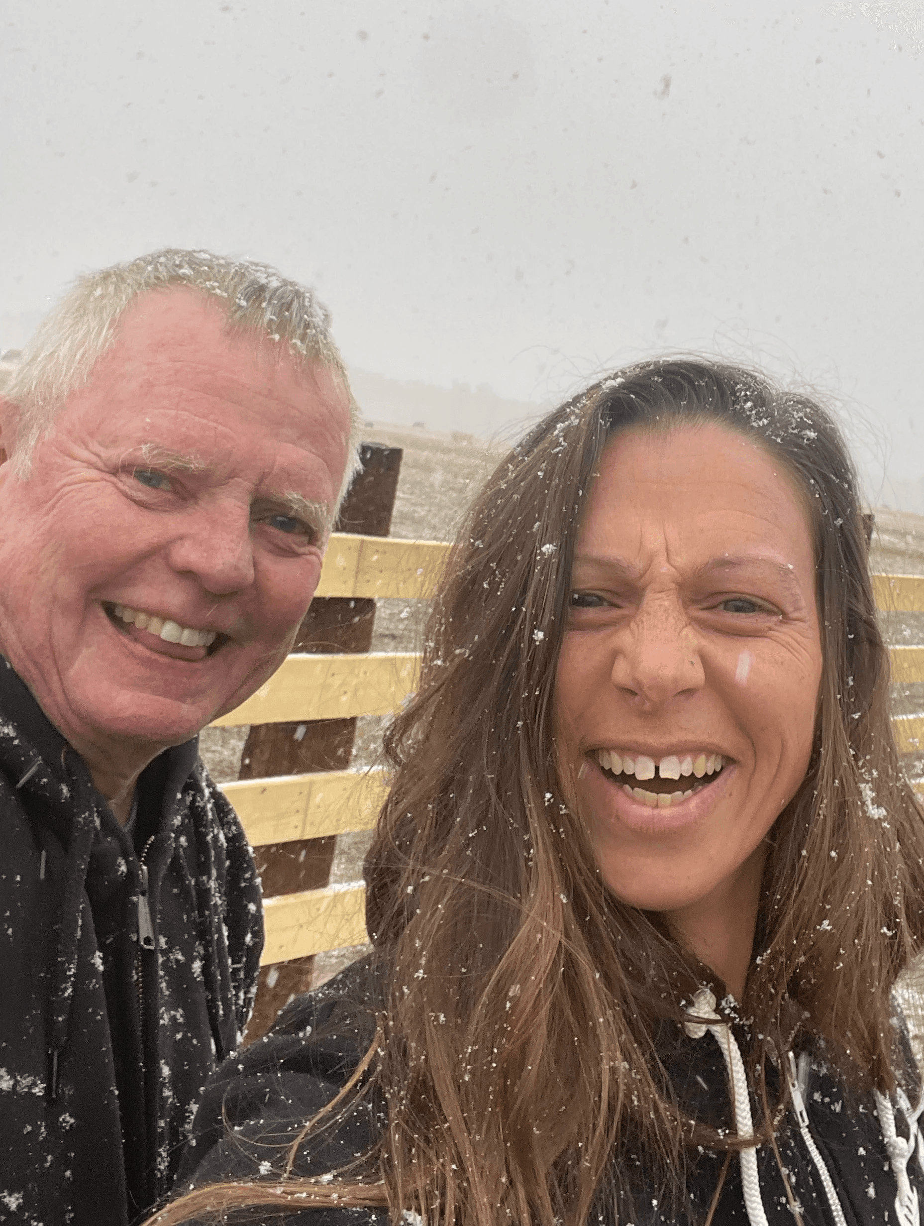 Two people smiling and posing for a selfie outdoors, with a blurred background of a building and cloudy sky.